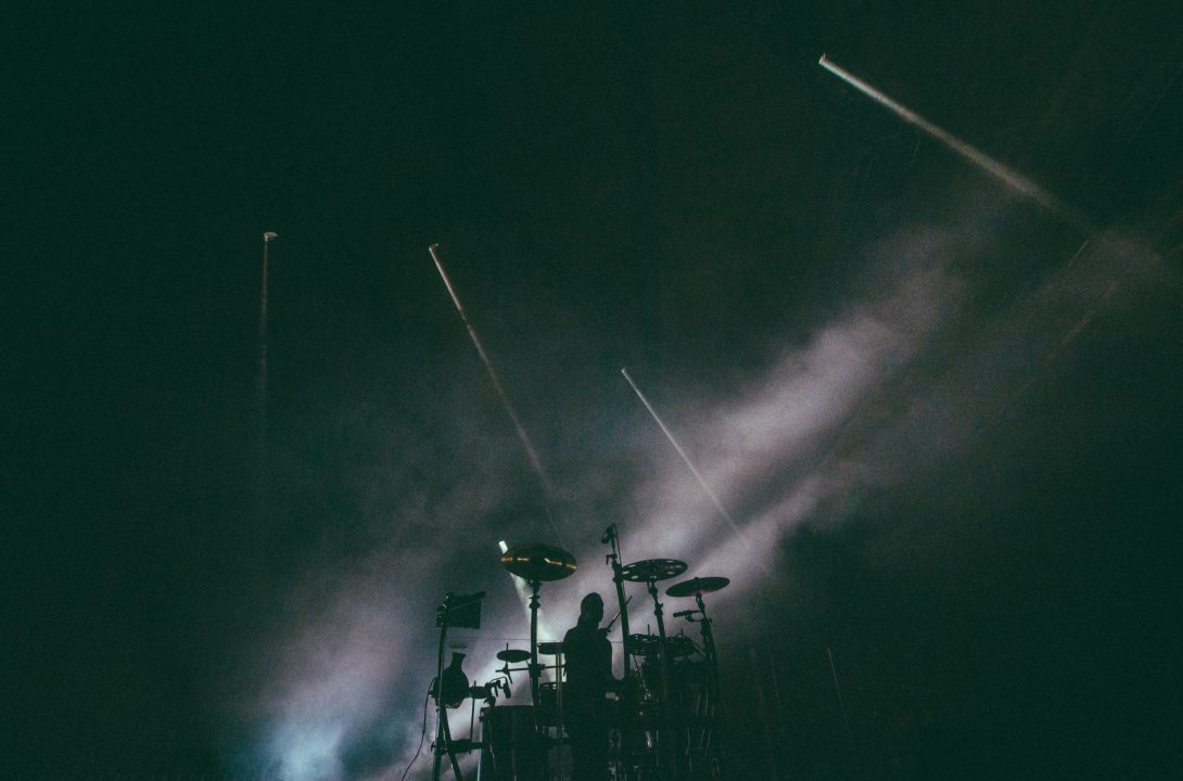 Daniel de los Reyes silhouetted at his percussion rig beneath sweeping stage lights